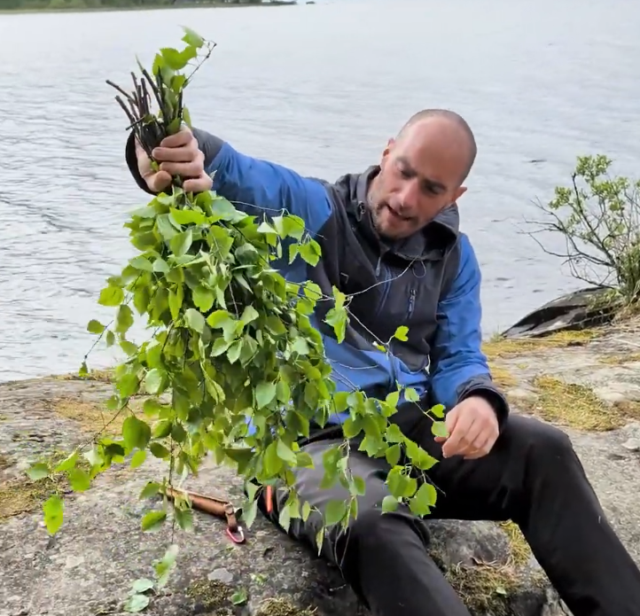 Stefano Mini prepara un fascio di rami di betulla freschi per la sauna, seduto su rocce vicino a un lago.
