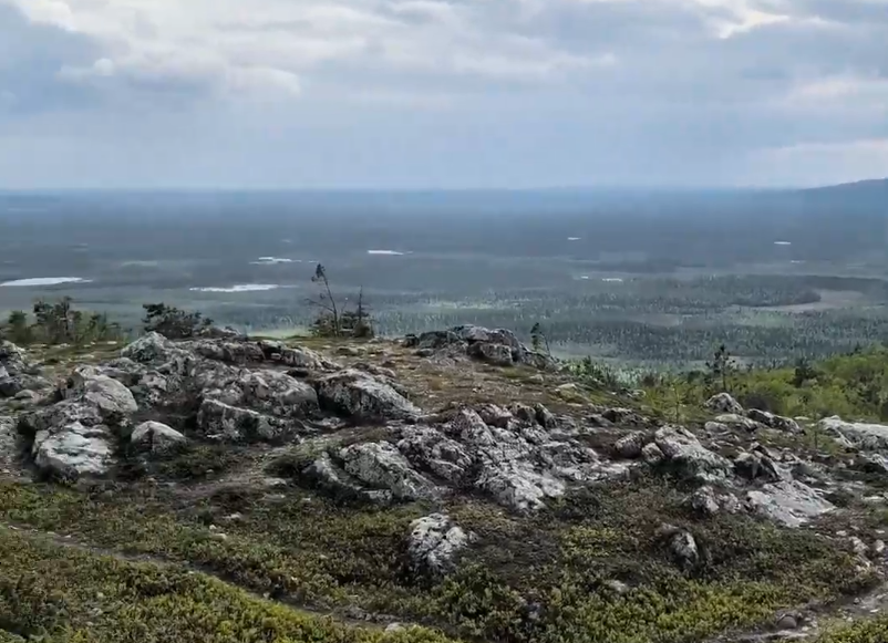 Vista panoramica di un paesaggio montano con rocce in primo piano, foreste e laghi sotto un cielo nuvoloso.