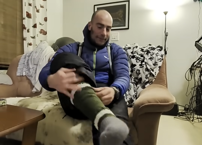 Stefano Mini si siede su una poltrona e si mette un calzino verde a strisce bianche. Indossa una giacca blu e bretelle.