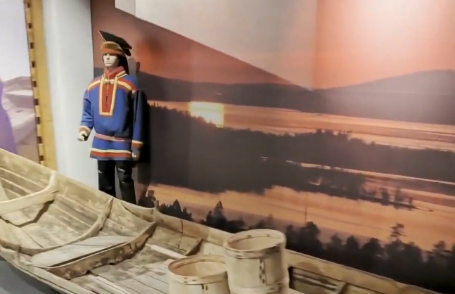 Manichino in abito tradizionale Sami blu e rosso, con cappello, accanto a una barca di legno. Sfondo di paesaggio con tramont