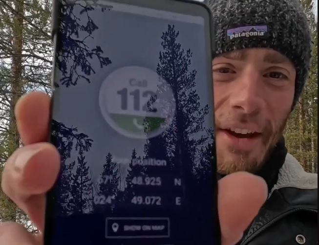 Stefano Mini mostra lo schermo del telefono con la chiamata di emergenza al 112 e le coordinate GPS in un bosco innevato.