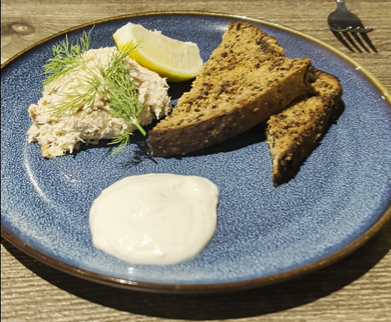 Pâté di pesce con aneto, limone, pane tostato scuro e salsa bianca su piatto blu, con forchetta sullo sfondo.