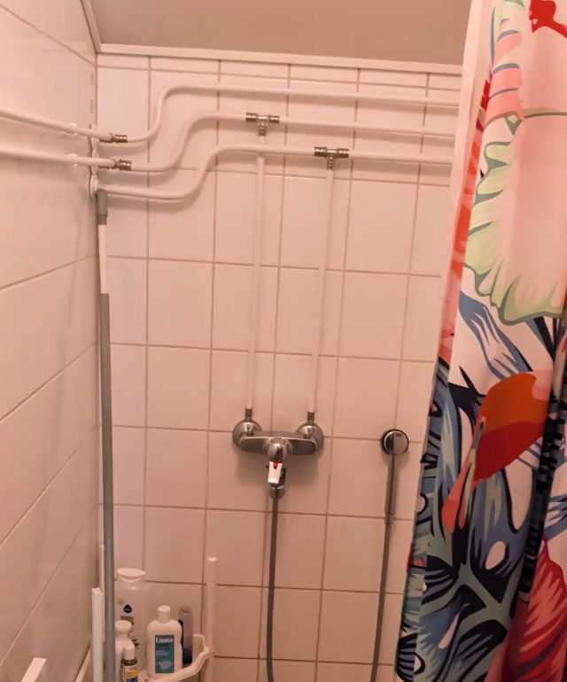 Doccia con piastrelle bianche, tubi a vista, miscelatore e tenda colorata con motivi floreali.