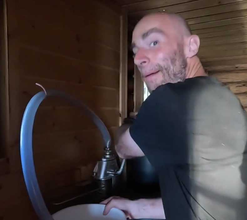 Stefano Mini sorride mentre usa un tubo per riempire un lavandino in una stanza di legno.