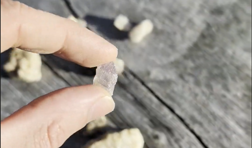 Mano che tiene un piccolo frammento di cristallo viola chiaro su una superficie di legno sfocata.