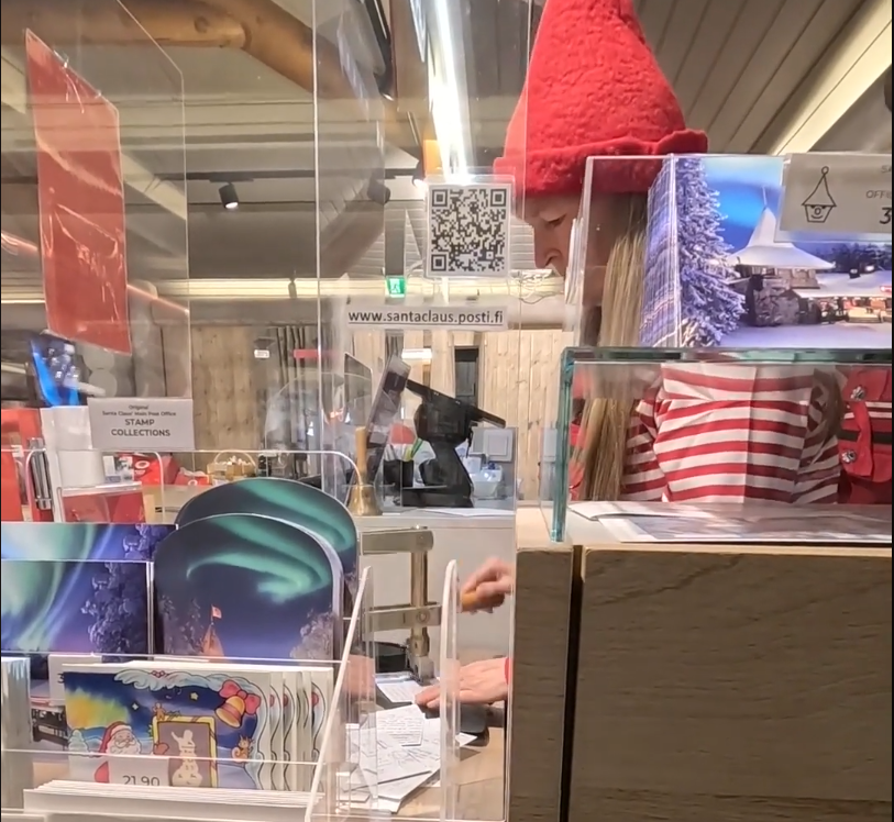 Donna con cappello da elfo rosso e maglia a righe bianche e rosse dietro un bancone con cartoline e QR code al Santa Claus Po