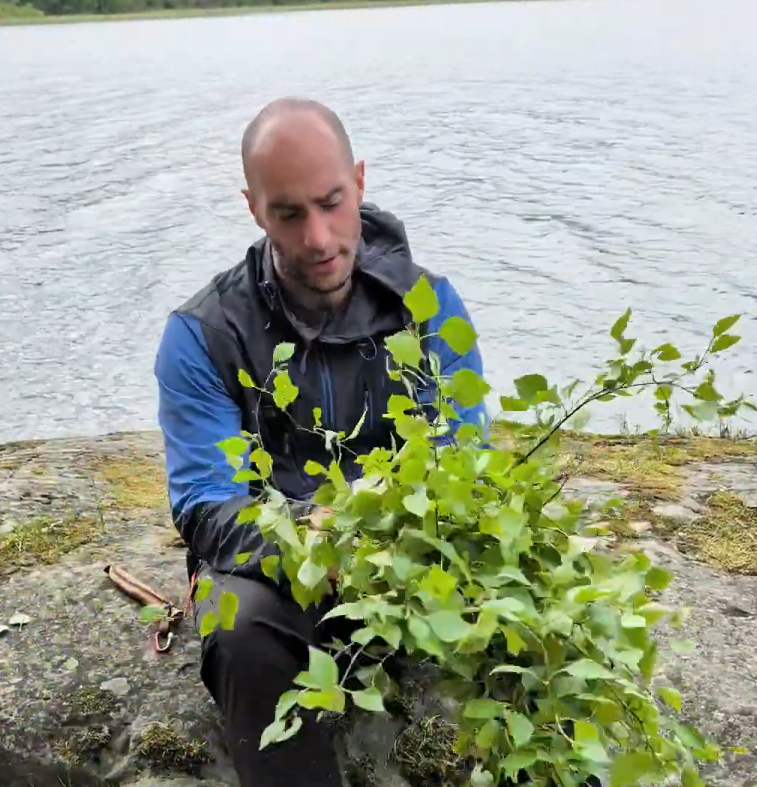 Stefano Mini siede su una roccia vicino all'acqua, tenendo in mano un fascio di rami verdi appena tagliati con delle cesoie a