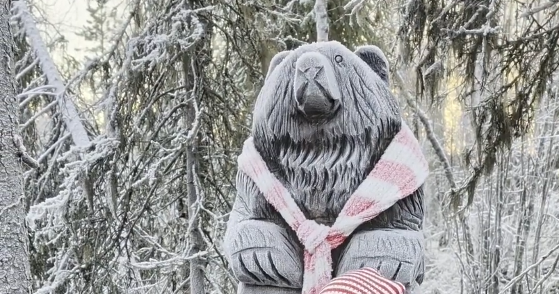 Statua di orso in legno con sciarpa a strisce rosa e bianca in una foresta innevata, alberi coperti di brina.
